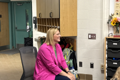 Woman in pink shirt sitting on chair in classroom, smiling at camera, surrounded by children.
