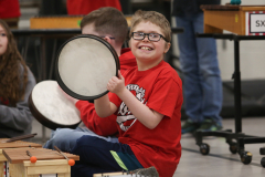 Boy in red shirt holding a drumstick in his hand, playing a drum.