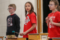 Three children playing musical instruments in a school setting. The child on the left is holding a blue mallet and wearing a