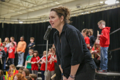 Woman standing at podium addressing audience of children in school auditorium.