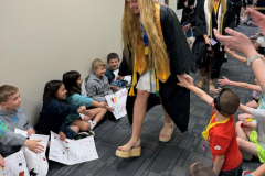 Woman in graduation gown walking through a crowd of children, some holding signs, others clapping and cheering.