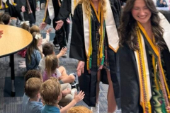 Graduates in caps and gowns walking on stage, children reaching out to touch them, a group of people clapping.