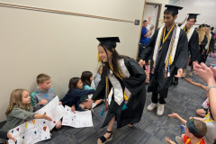 Graduates in caps and gowns walking down a hallway, children reaching out to them, people clapping, signs of celebration,