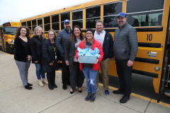 Group of people posing in front of a school bus; one person holding a blue gift bag.