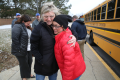 Two women hugging in front of a school bus, one wearing a red coat and the other a black beanie, posing for a picture.