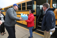 Two people interacting in front of a school bus, one holding a gift box, another handing it over.