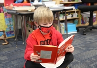 Child in a red jacket sitting on floor reading book.