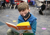 Young boy wearing a mask reading a book in a classroom setting.