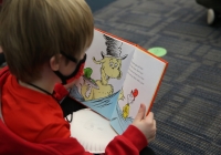 Child reading a book on Dr. Seuss Day, wearing glasses and a red jacket.