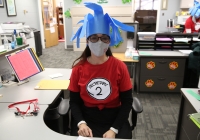Woman in office chair wearing blue bird costume, red t-shirt with comic strip character, and mask.