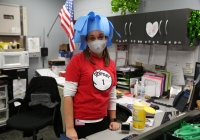 Woman in red shirt and blue hat stands at desk in office, celebrating Dr. Seuss Day.