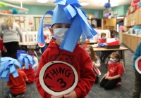 Child in a classroom on Dr. Seuss day, wearing a mask and holding a sign that says "Thing 3".