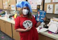 Woman in a Dr. Seuss costume holding a book titled "The Cat in the Hat" in a classroom setting.