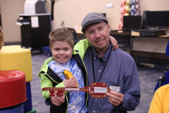 Man holding a young boy in front of a table with a red ribbon on it. The man is smiling and wearing a blue shirt.