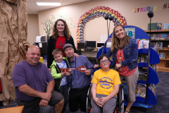 The group of people posing in front of a colorful display at a library event, celebrating Elliot's new role as mayor of