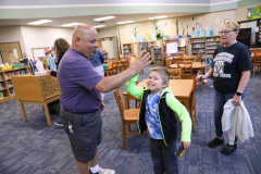 Man in blue shirt giving a high five to a young boy. They are standing in a library with other people around them.