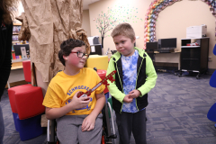 Two boys in a library, one wearing glasses, standing next to each other, looking at something off-camera.