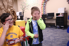 Three children in a classroom setting, one of whom is holding a guitar, all looking at something off-camera with expressions