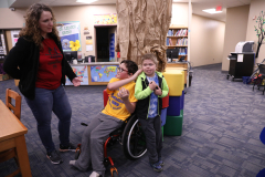 This is a photograph of a woman standing in a library, smiling at a young boy seated in a wheelchair. The boy is holding a