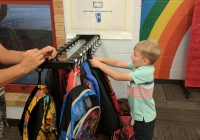 Young boy reaching out to grab a backpack from a rack.