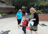 Three women walking on a sidewalk with a young boy, all looking down at papers. The woman in the middle is wearing a black