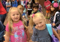 Two young girls standing next to each other, smiling at the camera, wearing backpacks and holding books, on a school day.