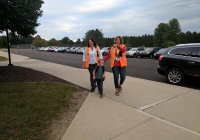 Two women walking on sidewalk near parking lot filled with cars. One woman holding child's hand. Both wearing safety vests.