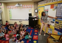 Teacher in front of whiteboard with children seated on floor behind her, pointing to chart.