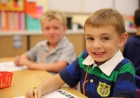 Two young boys sitting at a desk in a classroom, one of them is smiling and writing on paper.