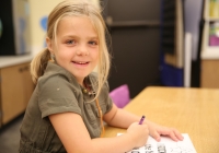 Young girl sitting at desk in classroom, smiling and looking towards camera, holding pencil over paper, first day of