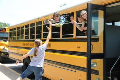 A school bus parked at a stop with students boarding, one person waving from window, last day of school 2023-24.