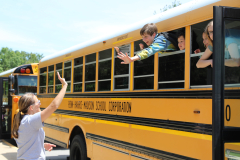 A yellow school bus filled with students waving goodbye to a woman standing outside.