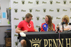 Two people sitting at a table with a microphone, in front of a banner that reads "PENNHILL" and has a logo for "Fighting