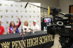 People seated at a table in front of a camera, microphone, and sign for Penn High School, with a woman standing behind them
