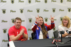 Three people posing at a press conference. A woman holding up a peace sign, a man clapping, and another person sitting