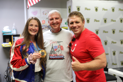 Two people posing with a trophy, one person wearing a red shirt, another in blue and white, and an American flag in