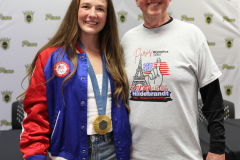 Woman in red, white, and blue jacket posing with older man wearing a medal around his neck.