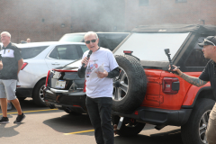 An elderly man speaking at an outdoor event, surrounded by a crowd of people and vehicles.