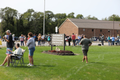 People gathered in a park for a parade, standing around a sign that says "Sarah Hildebrandt Parade".