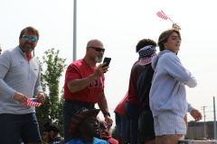 This is a photograph of an outdoor event where a group of people are gathered around a man holding a flag, and another