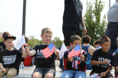 Children in sports jerseys holding flags, sitting on bleachers at a parade.