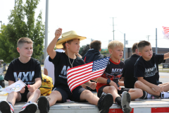 Boys in baseball uniforms sitting on a truck, holding American flags, celebrating at a parade.