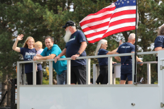 People on a parade float waving to crowd; American flag in background.