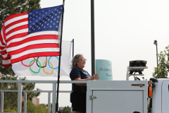 Person standing in front of a truck displaying an american flag, olympic rings, and a sign.