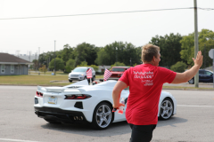 Man in red shirt waving at camera near sports car.