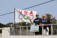Two people standing on a bridge holding a sign that reads "USA" and "Olympics". They are waving at the camera.