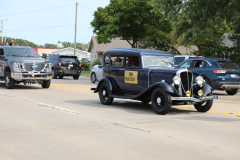 This is a photograph of an antique car driving on a street during a parade. The car has a yellow sign on its side that reads