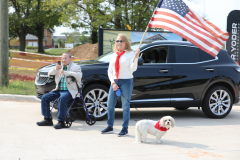 Two people standing next to a black car, holding an american flag. A dog sitting on the ground nearby.