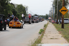 Parade of cars driving down a street in a small town.
