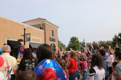 People gathered at a parade, waving flags and cheering, with a building in the background.